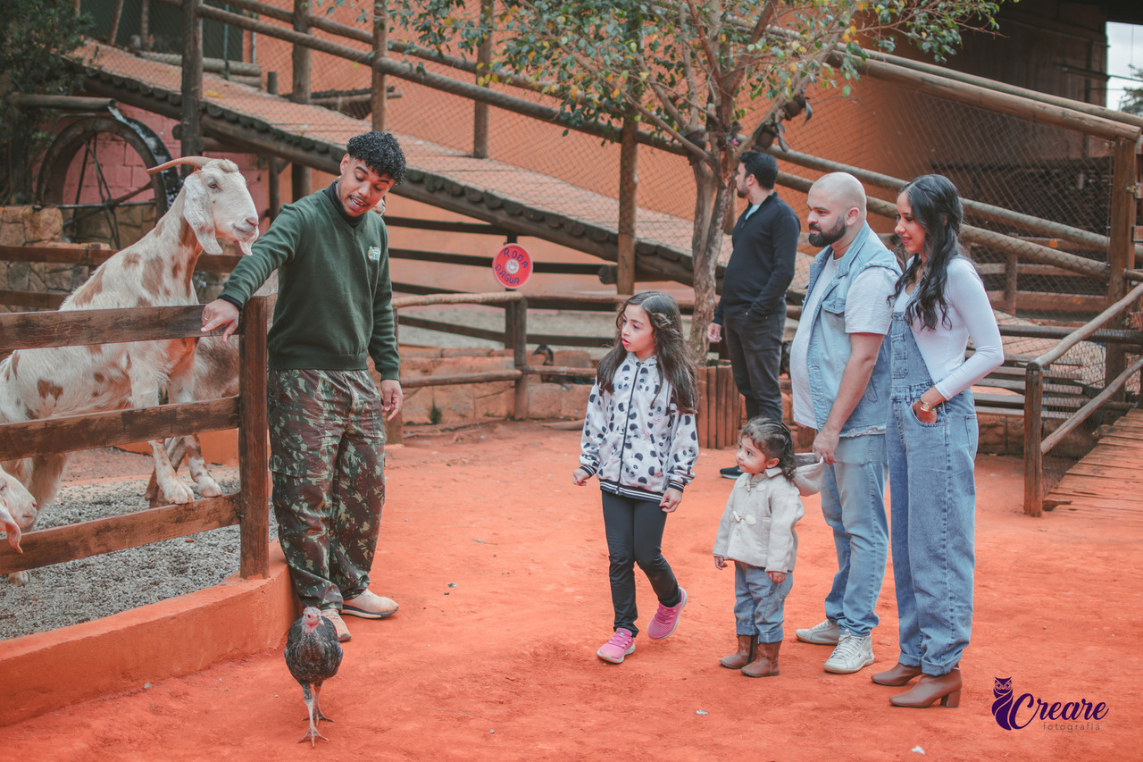 A imagem contém uma bode apoiado em uma cerca de madeira, um homem ao seu lado com calça camuflada e blusa verde. duas crianças, uma pequena com calça jeans e blusa de frio branca, outra criança maior com calça preta e blusa branca com pintinhas pretas. Uma mulher adulta com macacão jeans e blusa branca e um homem vestido de jeans. Todos observando o bode.