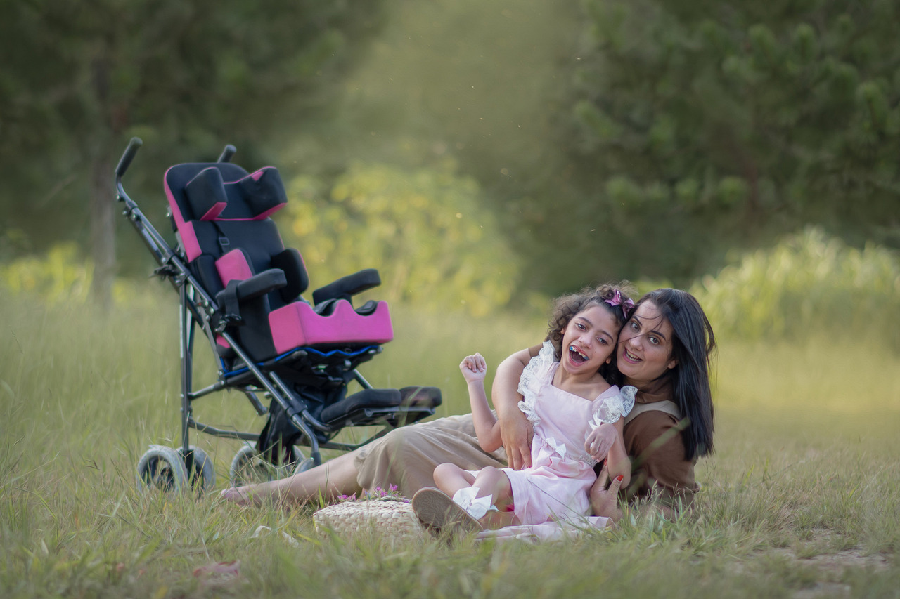 Uma mulher sentada na grama com sua filha, as duas dão risadas e ao lado a cadeira de rodas da menina que é PCD.