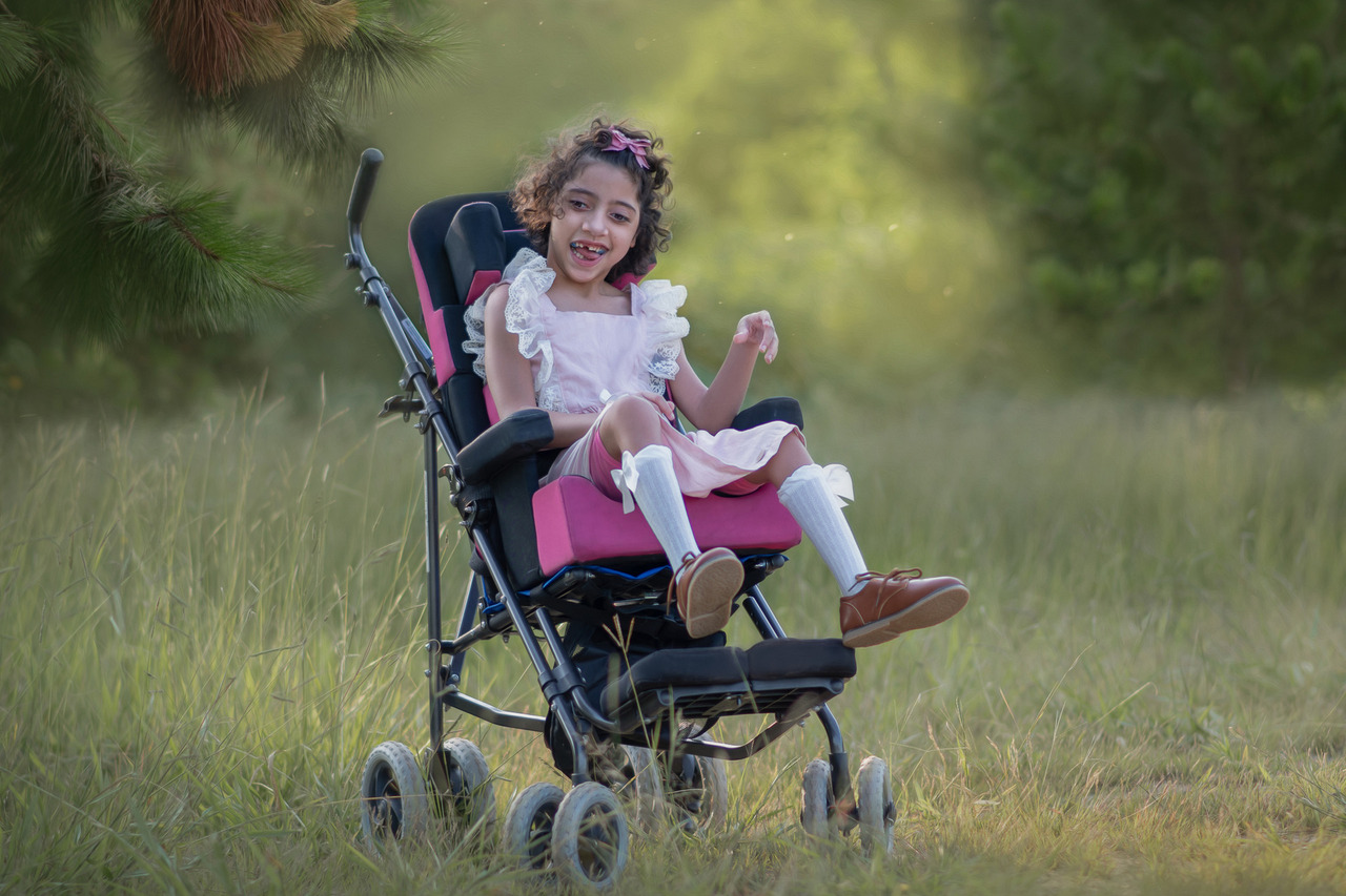 Uma menina cadeirante, sorrindo em um ambinete de natureza, durante ensaio fotográfico externo.
