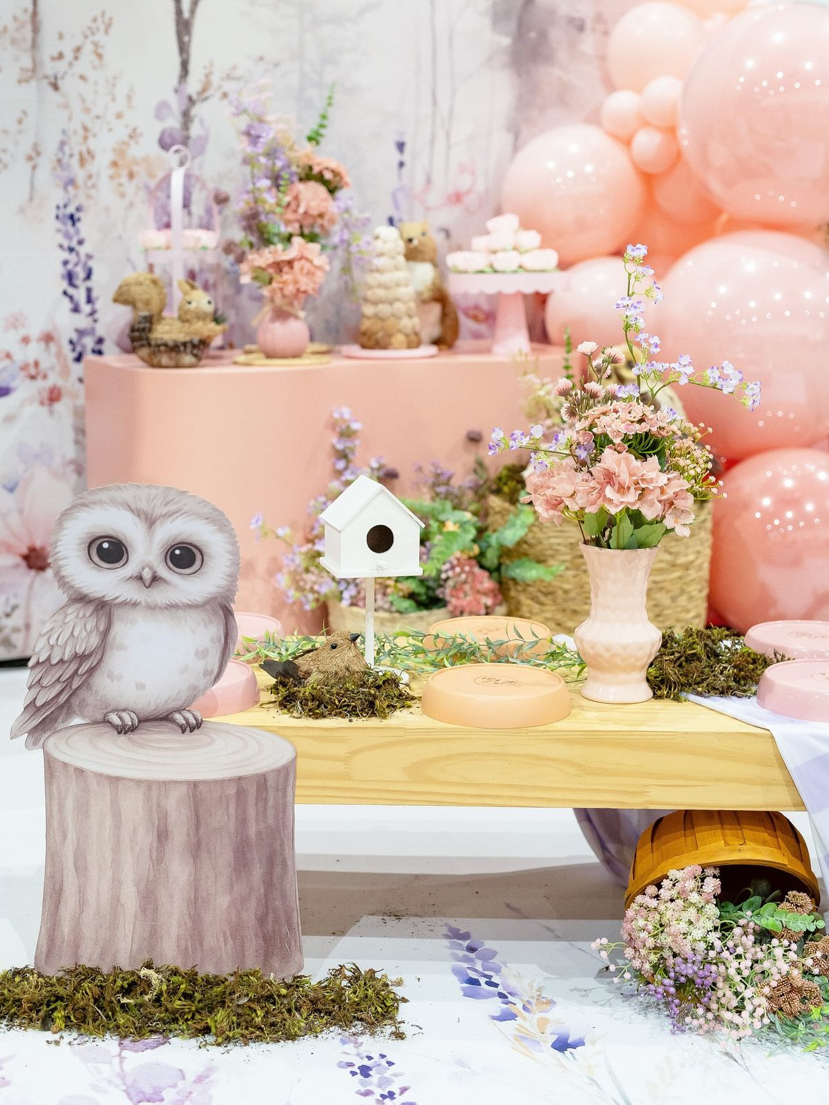 Decoração de festa infantil com tema coruja, mesa com flores uma pequena cas de passaro e balões rosa.