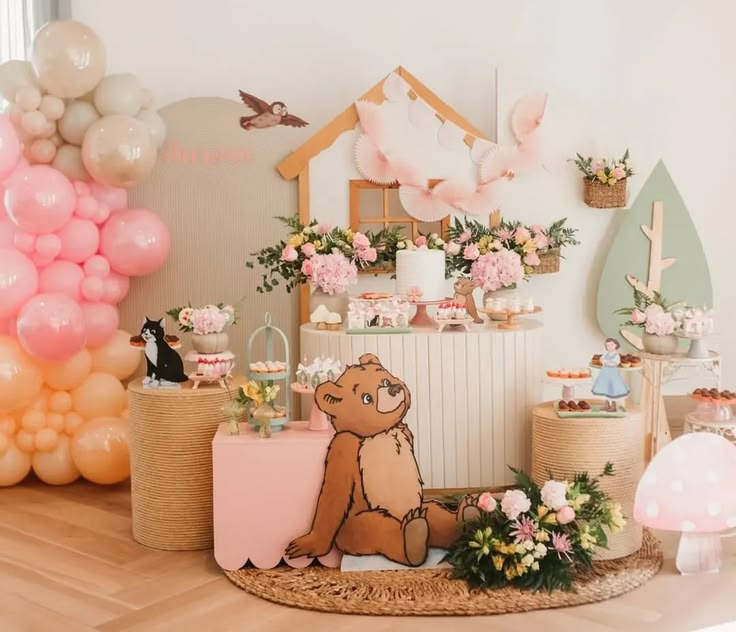 Decoração de aniversário infantil com tema de O Pequeno Urso, painel em tons pastéis com balões rosa e laranja e mesa com aranjo de flores.
