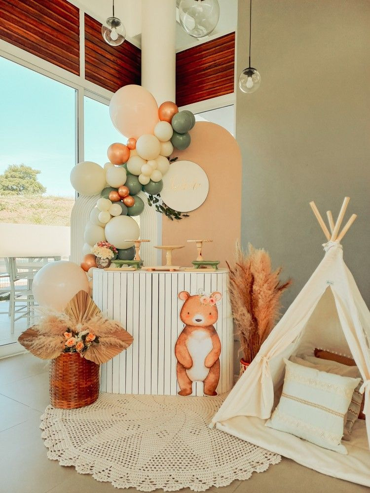 Decoração de aniversário infantil com tema acampamento, ao fundo balões em tons pastéis branco, verde e laranja, painel com um urso e flores e ao lado da mesma uma barraca de acampamento com almofadas brancas.