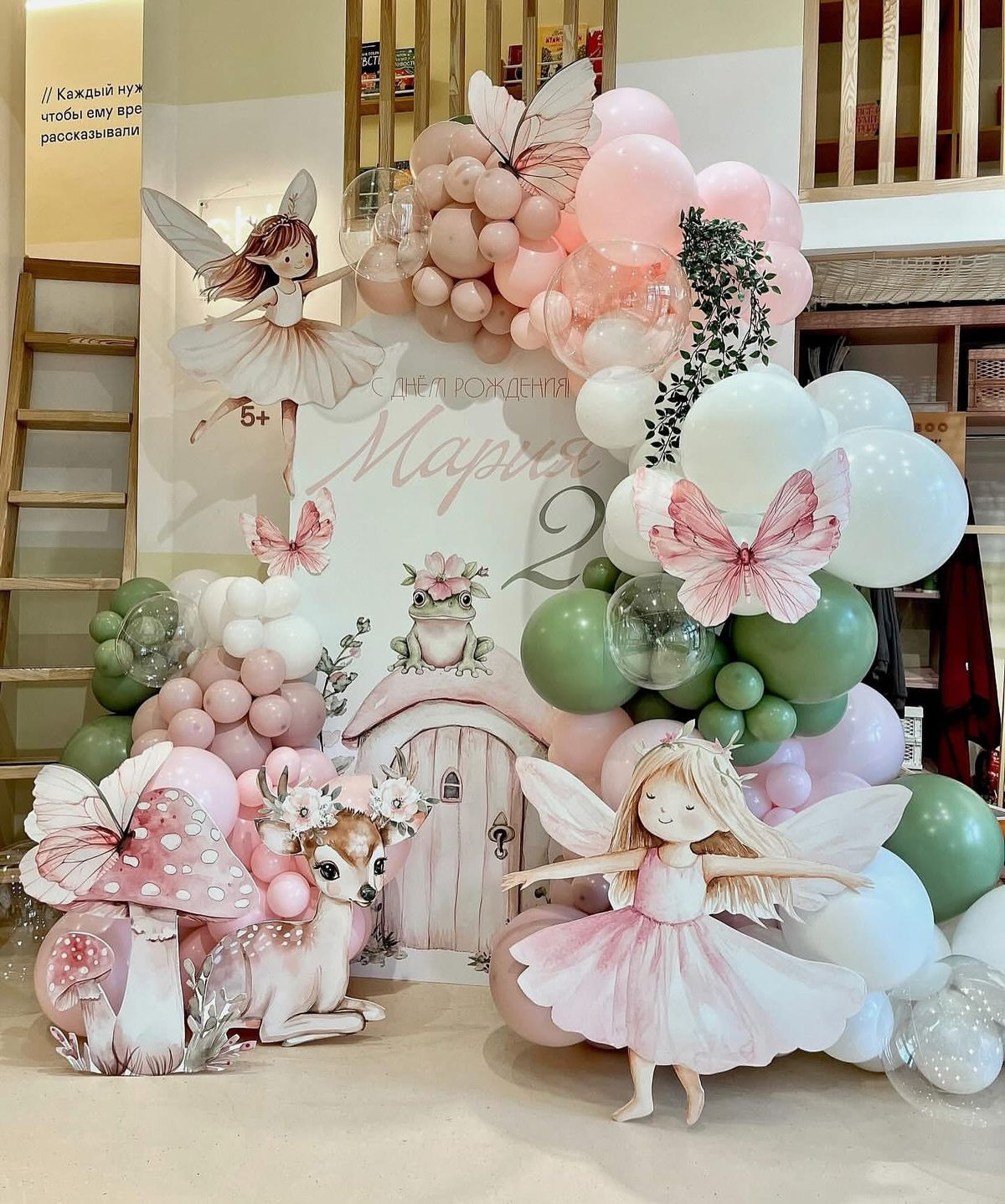 Decoração de festa infantil com tema jardim encantado, painel com fadas, cervo, cogumelos e casa mágica, arco de balões em tons pastel rosa, verde e branco.