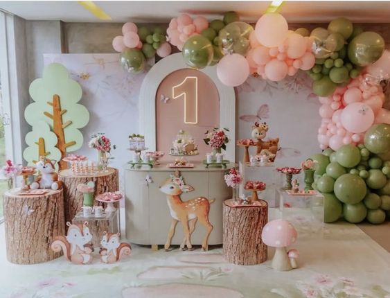 Decoração de aniversário infantil com tema floresta, a frente da mesa tem dois esquilos pequenos, um cervo filhote e cogumelos decorativos. Ao fundo balões rosa e verde na festa de 1 ano.