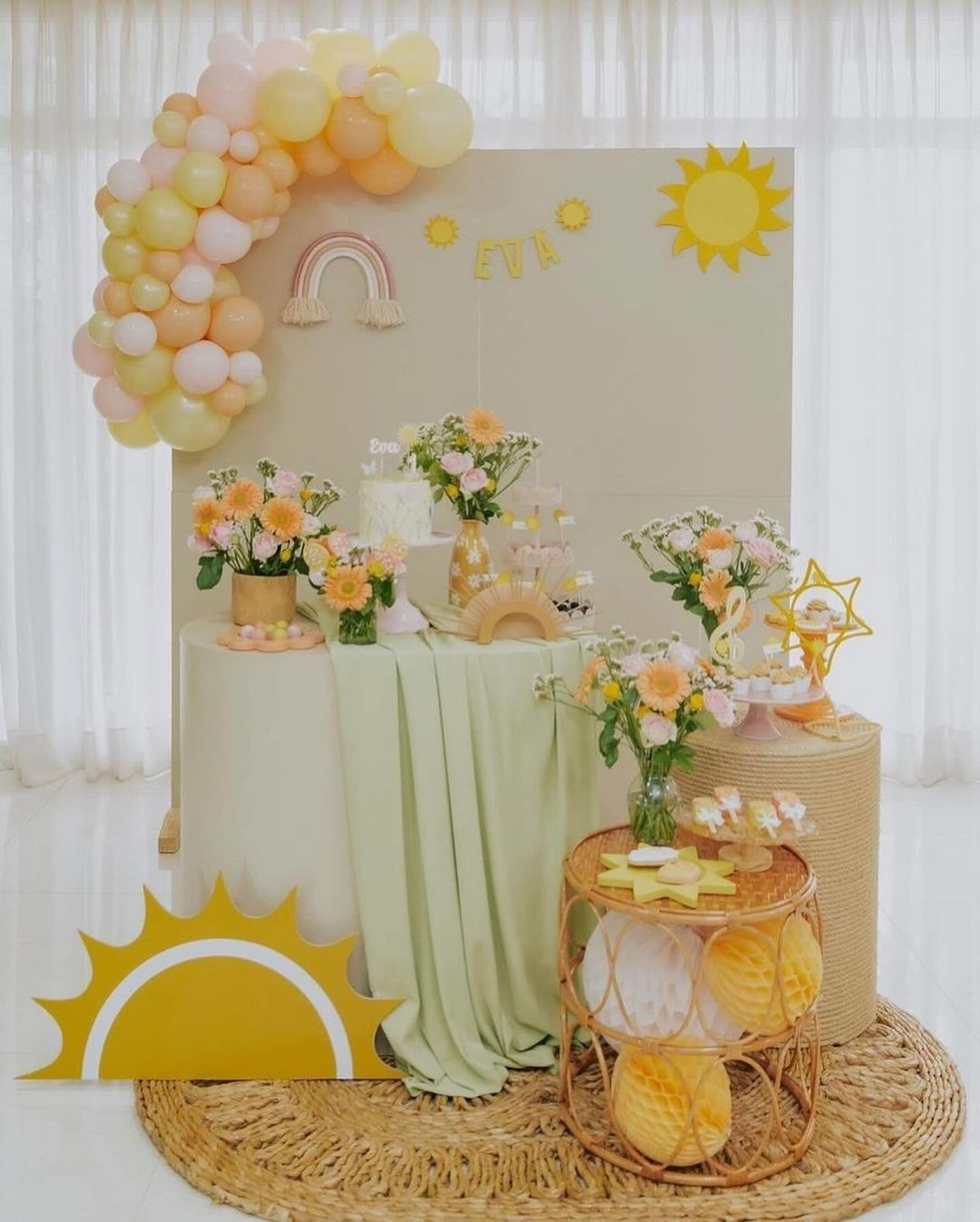 Decoração de festa infantil tema sol e flores, com arco de balões em tons pastel amarelo e verde e mesa delicada com arranjos florais.