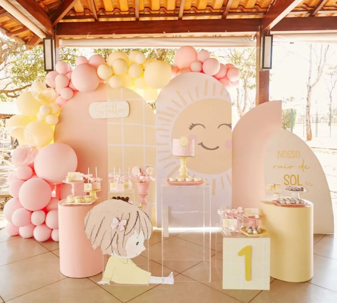 Decoração de festa infantil 1 ano tema Nosso Raio de Sol, com painel de sol sorridente, balões rosa e amarelo e mesa de doces personalizada.