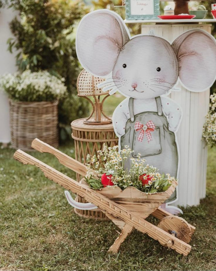 Decoração de festa infantil, painel com desenho de um rato, em tons delicados, vestido com uma jardineira verde e em frente um carrinho de madeira com flores.