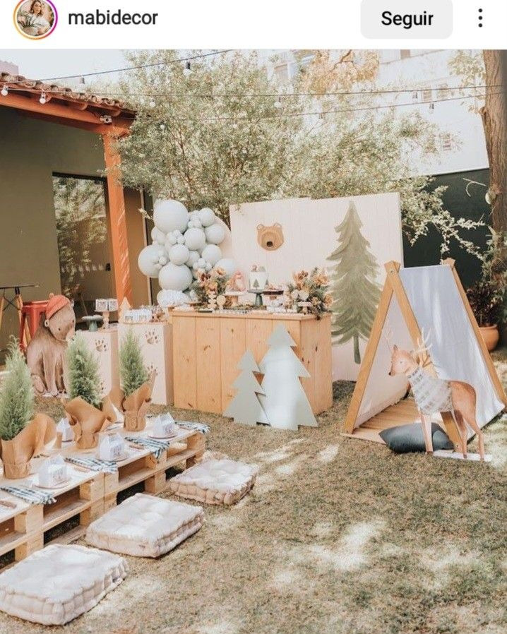 Decoração de aniversário infantil em casa, com tema Acampamento. Mesa decorativa com painel de arvores e uma barraca de camping ao lado, no jardim uma mesa com almofadas, para sentar .