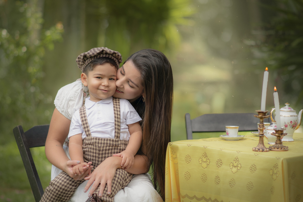 Uma mãe com seu filho no colo, sentados à mesa com toalha amarela preparada para um ensaio de dia das mães ao ar livre.