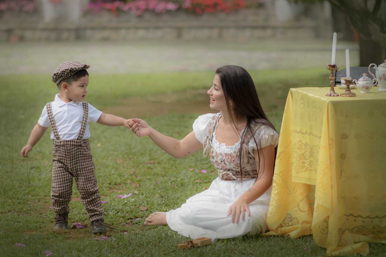 Mulher sentada na grama segurando a mão do filho que está em pé ao seu lado, próximos a uma mesa com toalha amarela preparada para um ensaio de dia das mães.