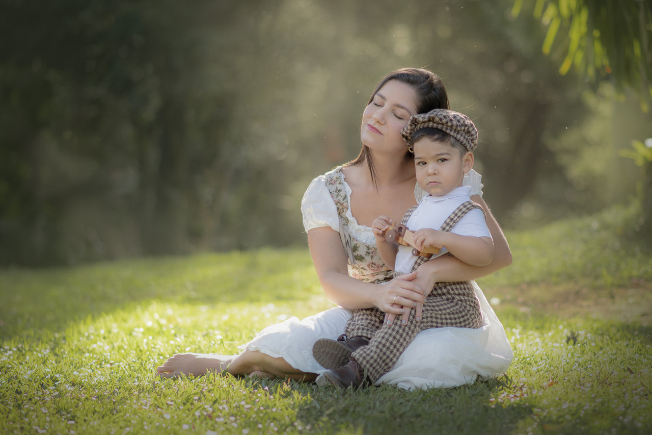 Uma mulher sentada na grama com seu filho no colo, com uma luz de fim de tarde envolvendo os dois, durante o ensaio de dia das mães ao ar livre.