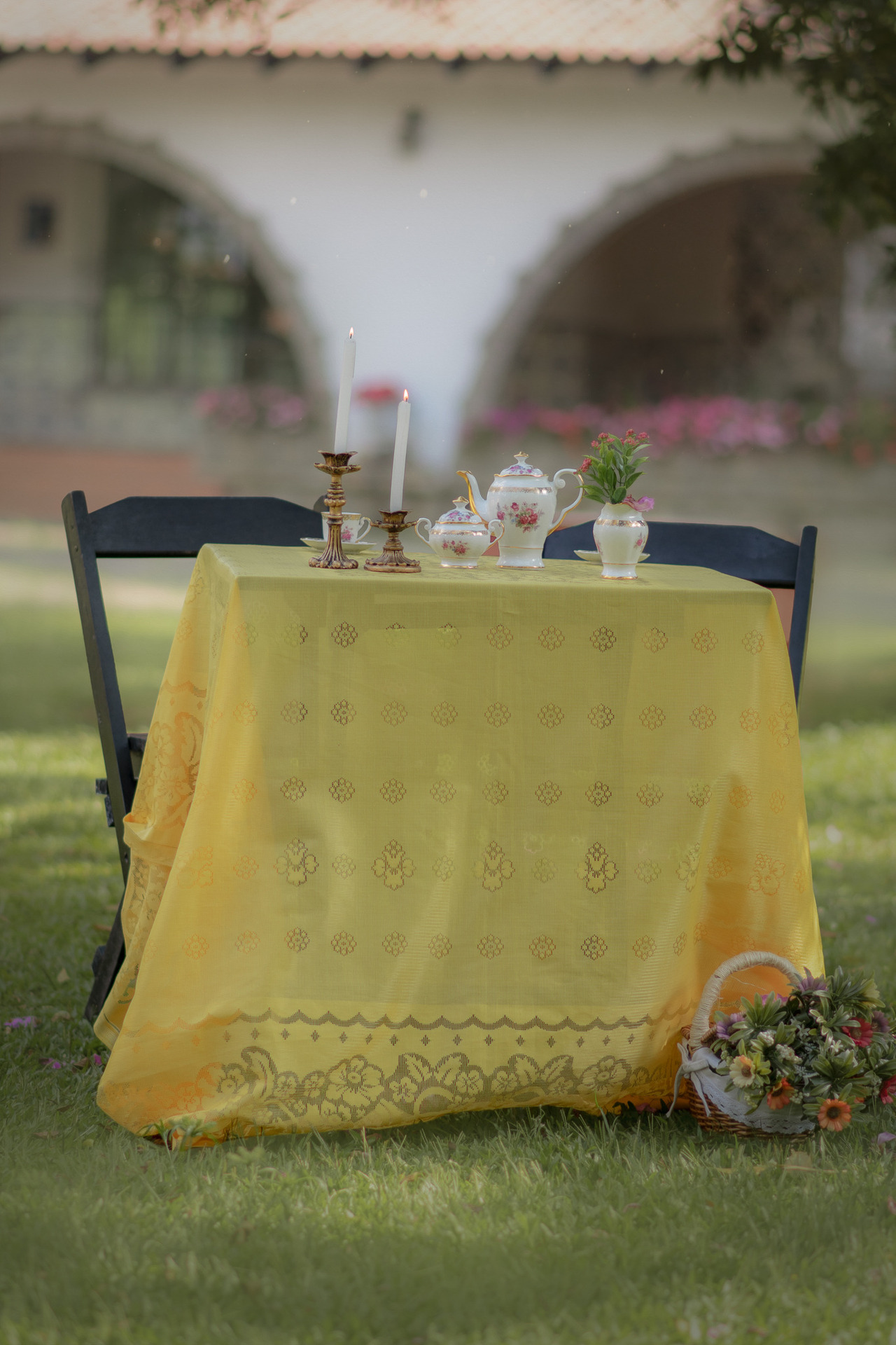 Cenário de ensaio fotográfico de dia das mães ao ar livre com mesa de chá, toalha amarela e dois castiçais com velas.