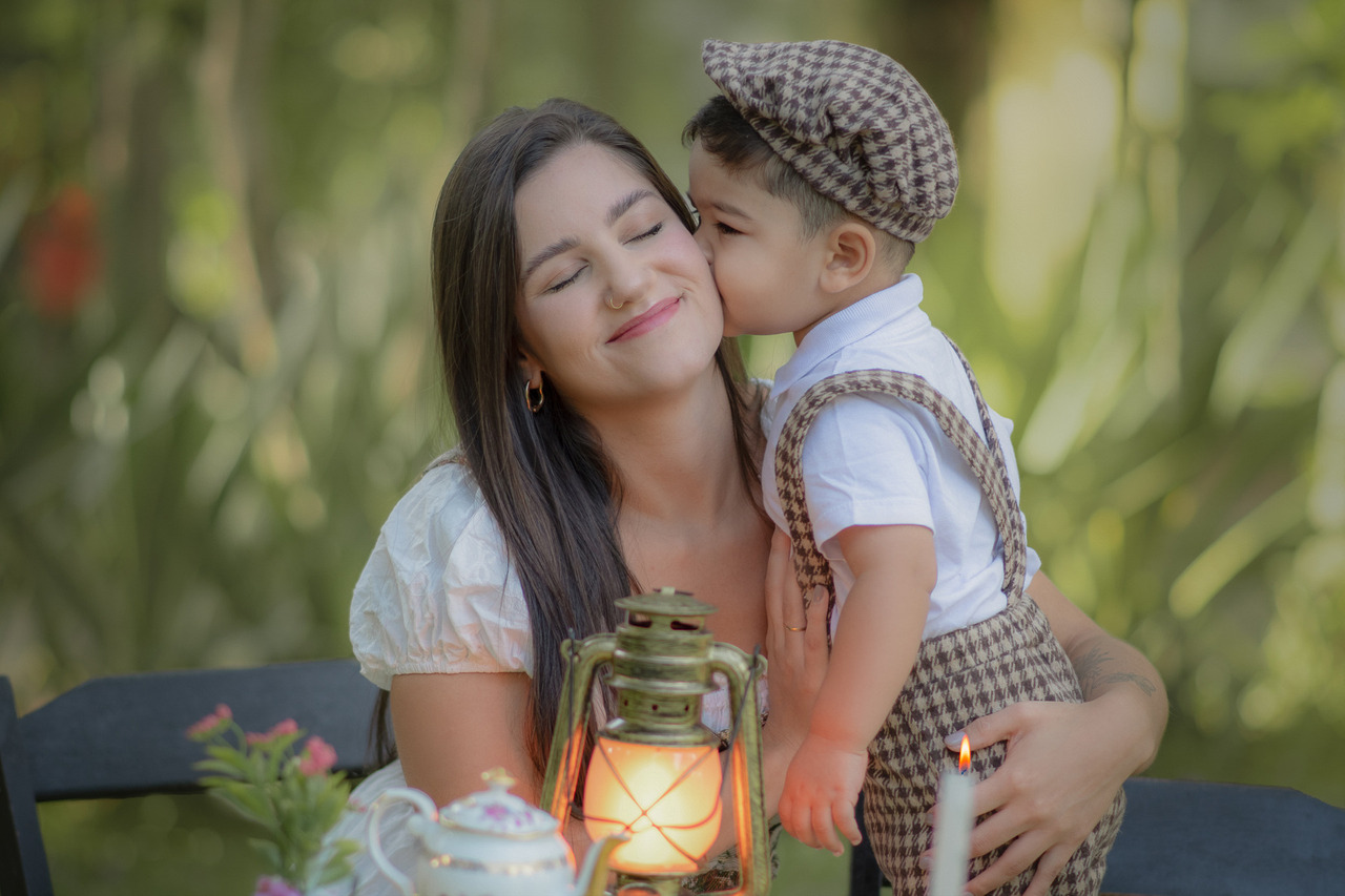 Mãe e filho sentados em uma messa em um parque, durante um ensaio fotográfico ao ar livre, o filho beija o rosto da mãe.