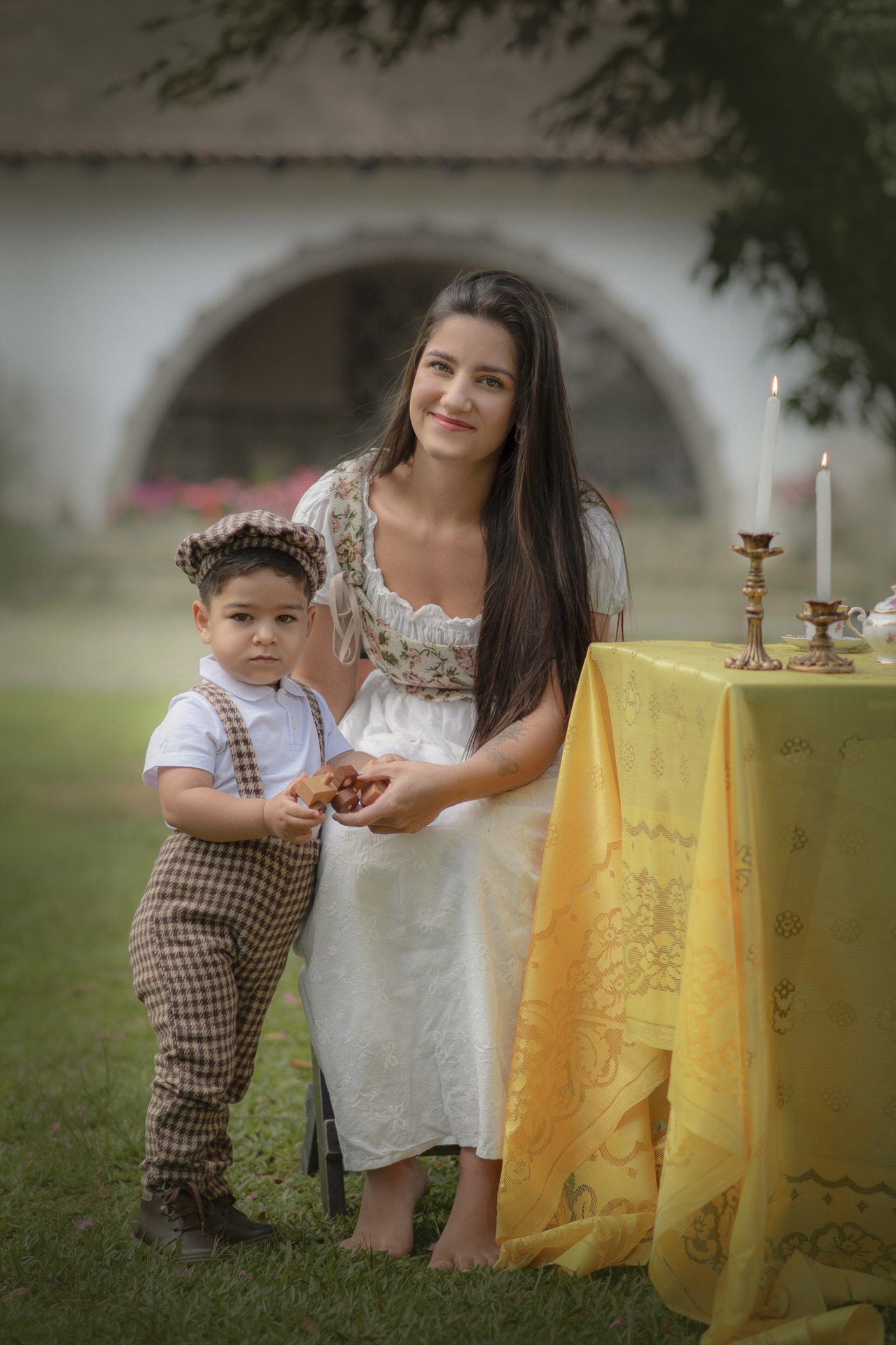 Uma mulher jovem usando um vestido branco e um corset, ao lado um menino vestido com roupas camponesas e uma boina, ao lado uma mesa com toalha amarela e castiçais de velas.