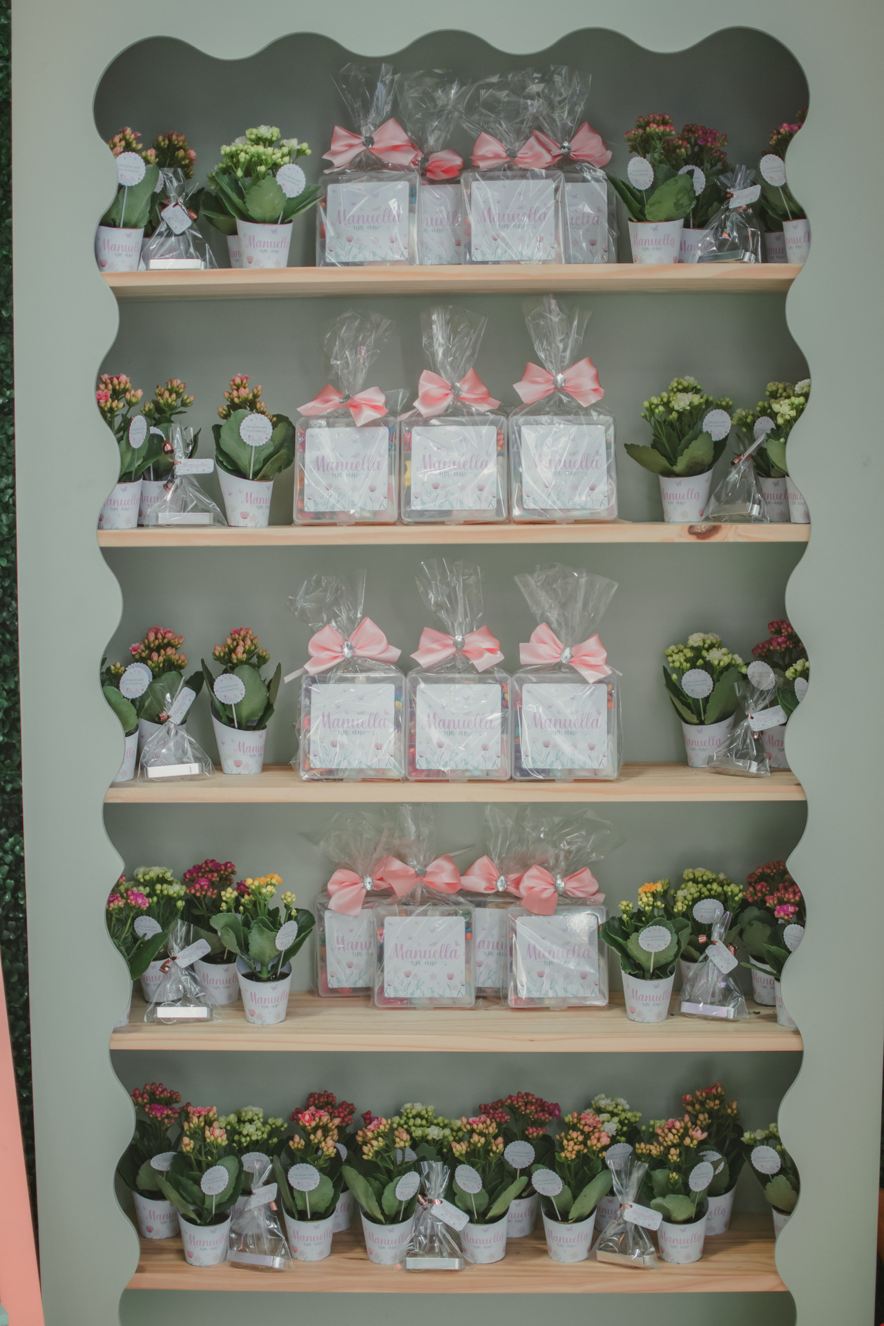 Lembrancinhas personalizadas para festa de 1 ano, organizadas em estante decorativa com laços rosa e vasos de flores.