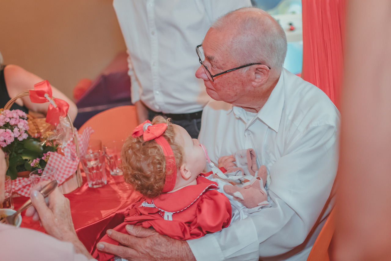 Uma bebê usando roupa vermelho no colo de seu bisavô. A bebê olha de forma amorosa para o senhor.