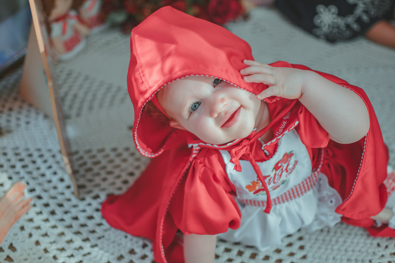 Uma bebê de um ano usando roupa de Chapéuzinho vermelho e sorrindo.