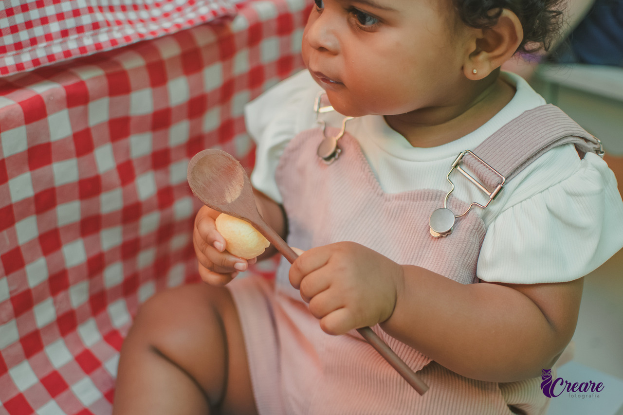 Uma bebê segurando uma colher de pau e experimentando um pão de queijo.