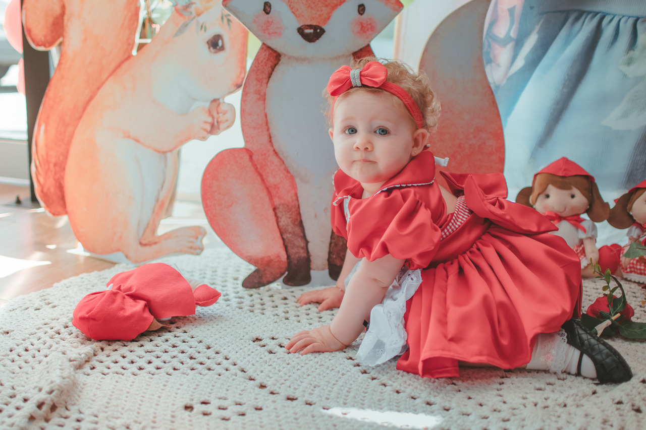 Uma bebê usando vestido vermelho e engatinhando em um tapete branco, durante seu aniversário de 1 ano.