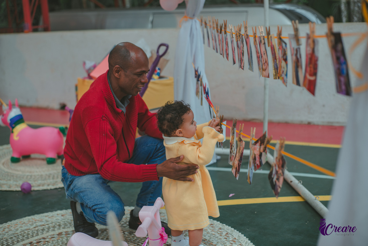Uma bebê usando vestido amarelo e um senhor usando blusa vermelha observando fotografia, durante festa de 1 ano.