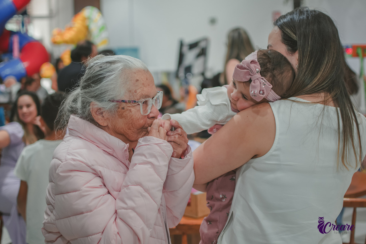 Uma senhora de idade beijando a mão de uma bebê, durante seu aniversário de 1 ano.