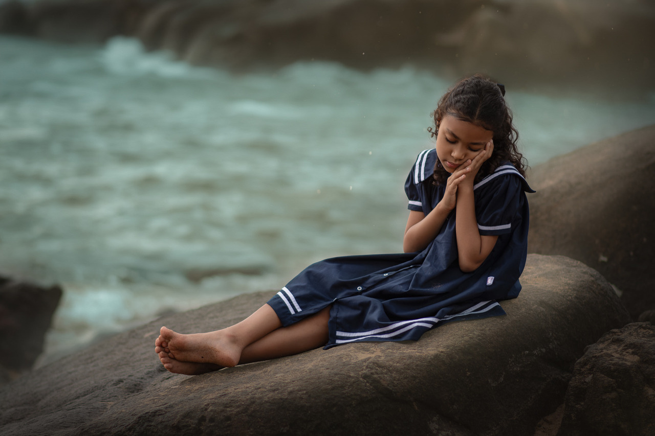uma menina usando vestido azul sentada em uma pedra na praia, durante ensaio fotográfico infantil.