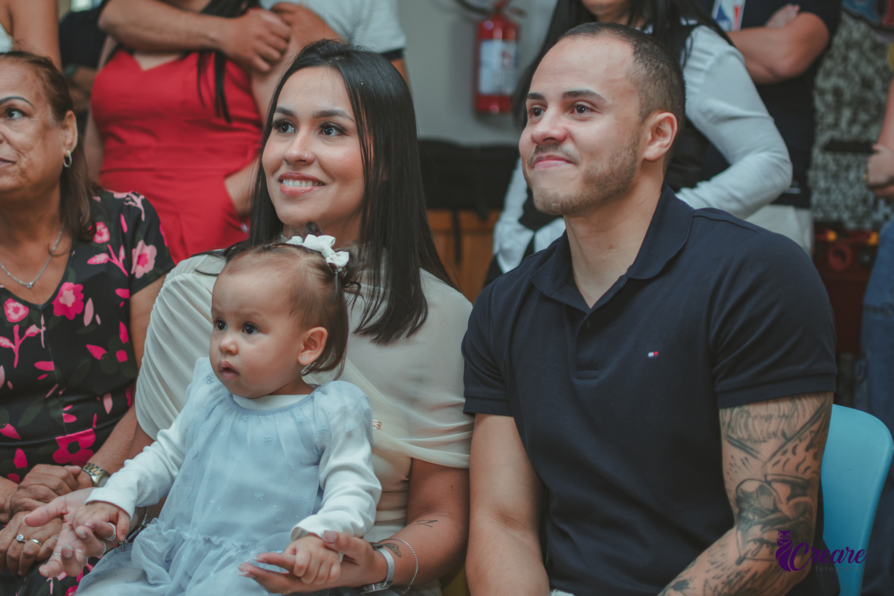 uma casal emocionado com sua filha de 1 ano no colo, enquanto assistem a retrospectiva da festa.