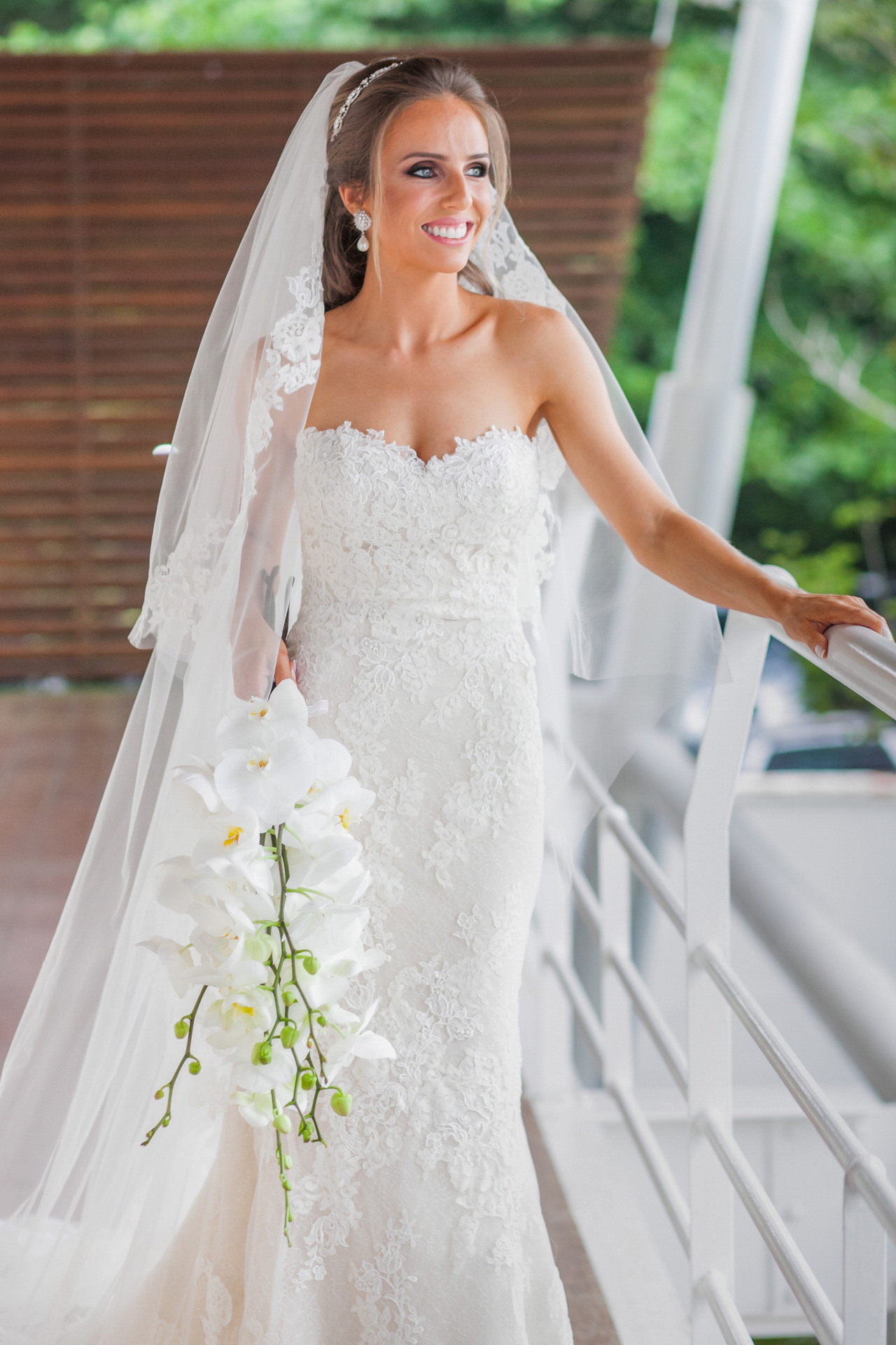 Uma noiva parada olhando para o lado segurando seu buque. pronta para o casamento. Ela tem olhos claros e cabelos castanhos esta usando um véu comprido e um vestido sem alças todo rendado em branco off white