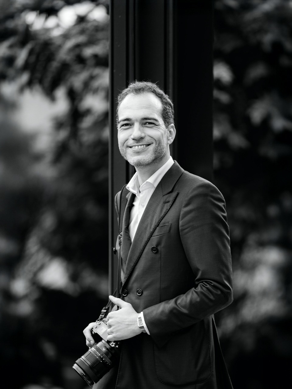 Filipe Santos vestido de fato e maquina fotografica na mão a sorrir em dia de casamento no jardim de uma quinta para casamentos