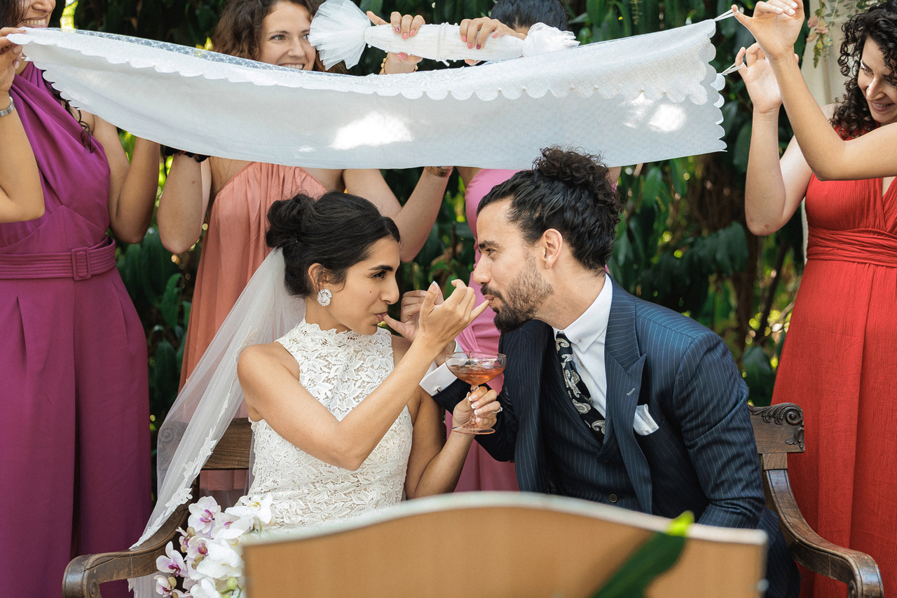 Noivos sentados a partilhar um momento descontraído, com amigas a segurar um pano branco suspenso sobre eles, num espaço exterior com vegetação. Foto por Filipe Santos, 2025.