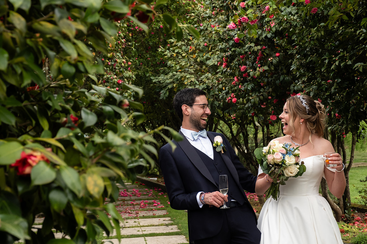 Noivos a caminhar e a rir juntos num jardim, ele com um copo na mão e ela com um ramo de flores, rodeados por árvores e vegetação. Foto por Filipe Santos, 2025.