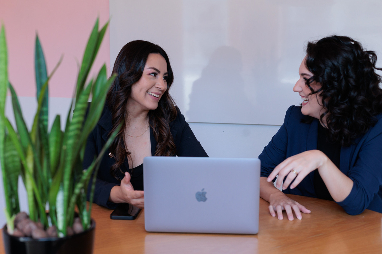 Retrato profissional de duas mulheres sorridentes conversando em uma reunião. Elas estão sentadas à mesa, com um laptop e um celular à frente, vestindo blazers escuros. O ambiente é moderno e descontraído, com uma planta decorativa em primeiro plano e uma parede em tons claros ao fundo.