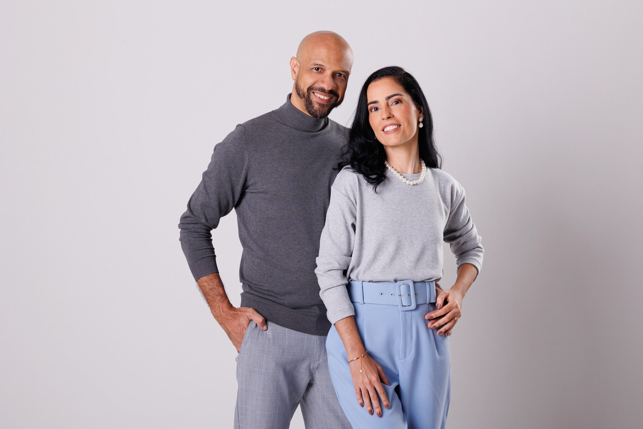 Retrato corporativo de um homem e uma mulher sorridentes, posando juntos em estúdio com fundo cinza. Ele veste suéter de gola alta cinza escuro e calça social; ela usa blusa cinza claro, calça azul clara e colar de pérolas, transmitindo elegância e profissionalismo.