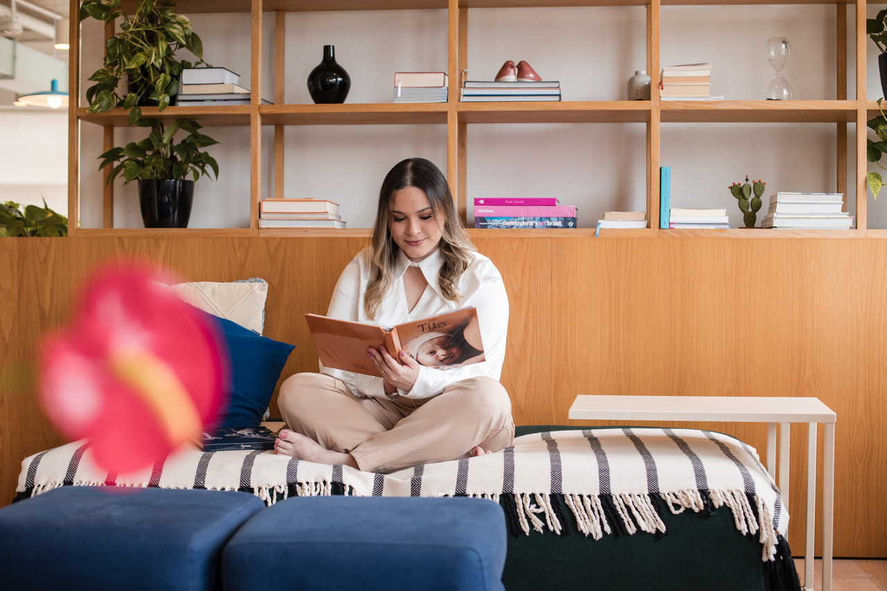 Retrato profissional de uma mulher sorridente, segurando dois álbuns fotográficos, sendo o da frente intitulado 'Ayanna', com a imagem de um recém-nascido na capa. Ela veste camisa branca e está sentada em um ambiente aconchegante, transmitindo alegria e orgulho pelo trabalho realizado.