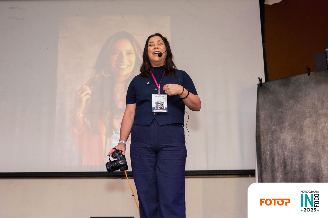 Ana Campbell palestrando no congresso de fotografia InFoco 2025 em Nova Iguaçu, especialista em retratos corporativos e fotografia para LinkedIn, segurando câmera fotográfica no palco durante sua apresentação sobre retratos de marca pessoal e posicionamento profissional.