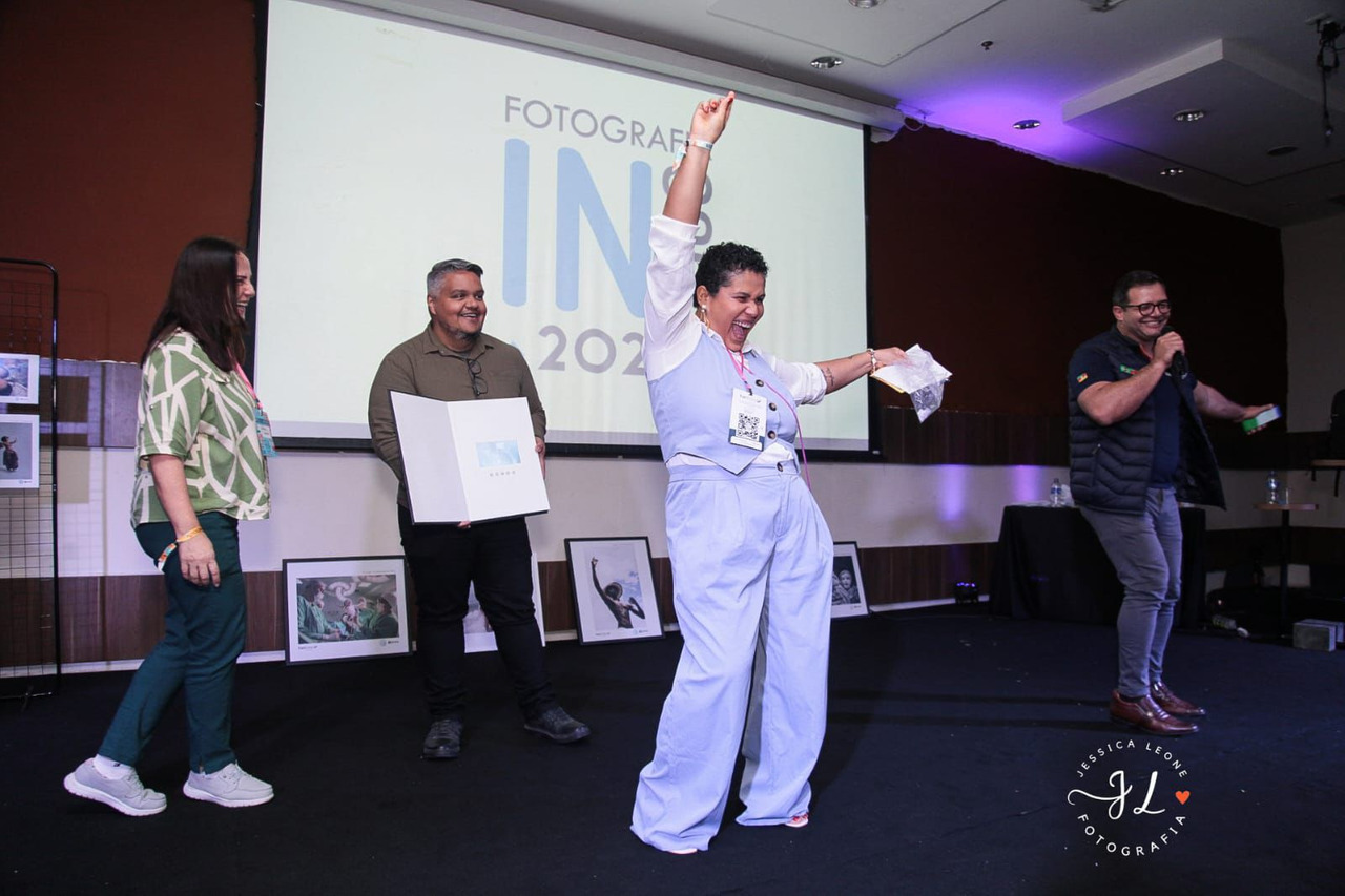 Premiação de fotografia no congresso Fotografia InFoco 2025: fotógrafa de terninho azul comemorando vitória no palco, de braços erguidos, enquanto outros participantes aplaudem. Evento de fotografia com destaque para premiação, fotógrafos e networking profissional.
