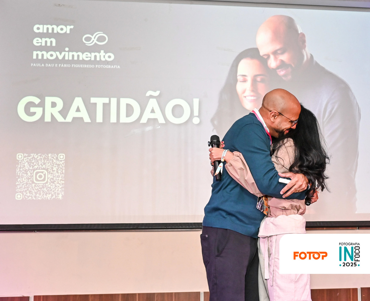 Casal de fotógrafos Paula Dau e Fábio Figueiredo se abraçam no palco do InFoco 2025, em frente à projeção com a frase 'Gratidão!' e o logotipo do projeto Amor em Movimento.