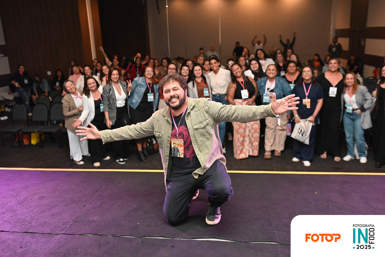 Palestrante no congresso de fotografia InFoco 2025 em Nova Iguaçu posa sorridente no palco com os braços abertos diante de plateia cheia de fotógrafos participantes do evento.