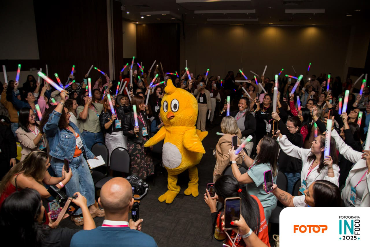 Interação divertida no congresso de fotografia InFoco 2025: mascote amarelo participa com a plateia que vibra com bastões de luz, em ambiente descontraído e cheio de energia no evento em Nova Iguaçu.