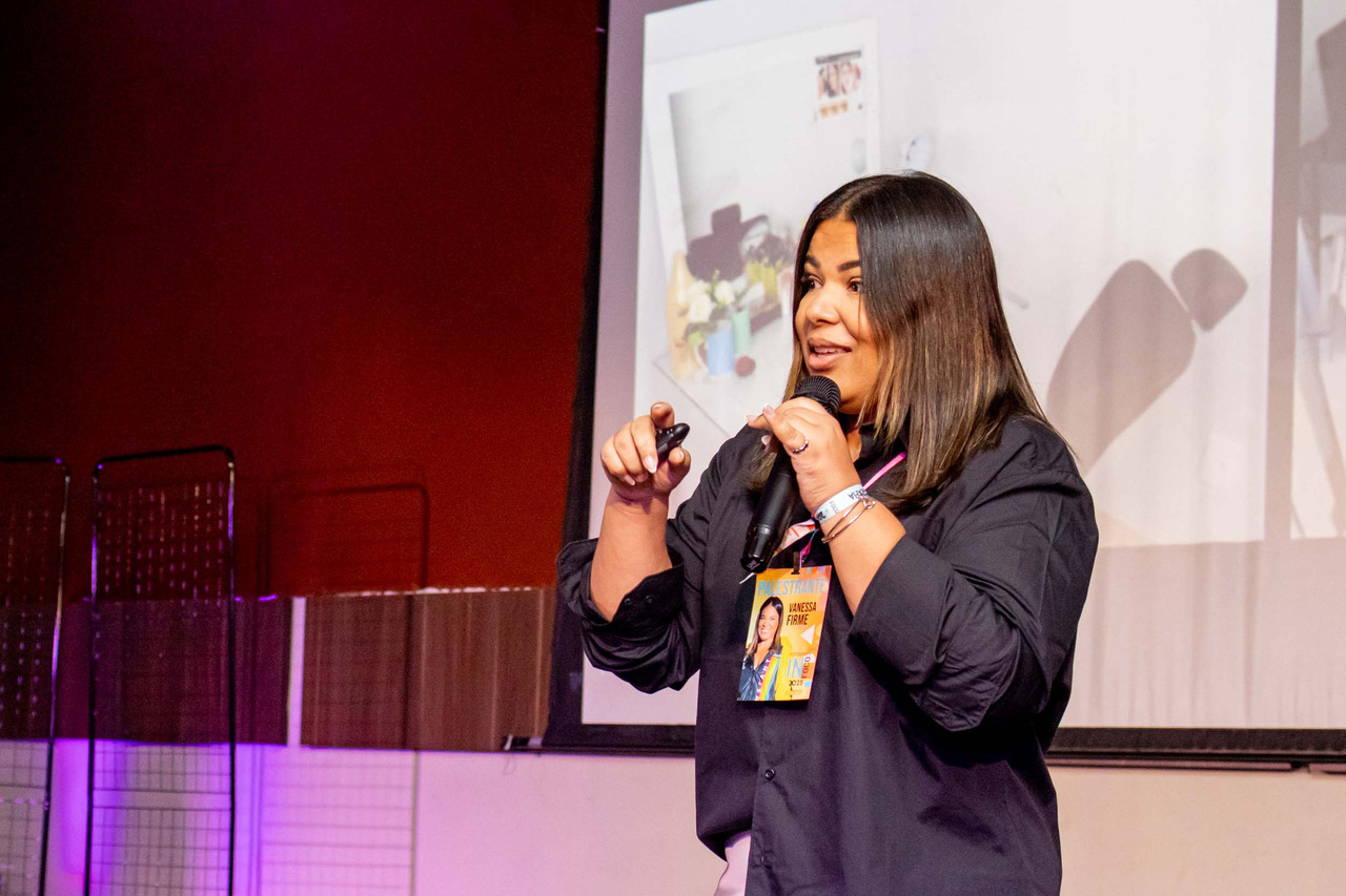 Vanessa Firme em palestra no InFoco 2025, congresso de fotografia em Nova Iguaçu, apresenta no palco com microfone e projeção ao fundo, compartilhando técnicas e experiências com fotógrafos.