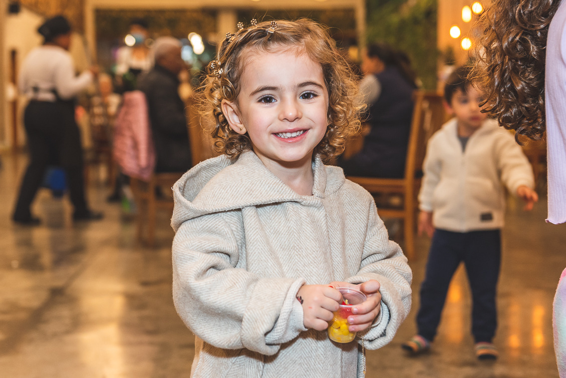 Menina sorrindo para a câmera durante uma festa infantil em ambiente interno, segurando um copinho de doces, com outros convidados desfocados ao fundo e luzes quentes decorativas.