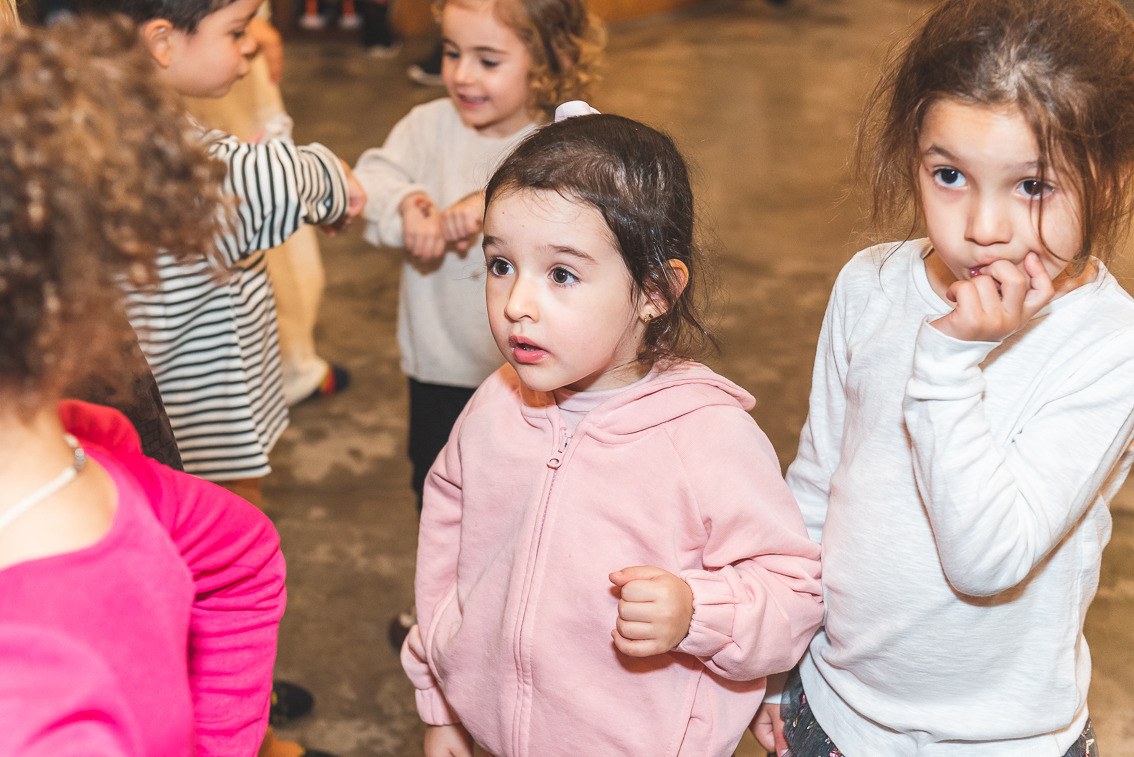 Grupo de crianças reunidas durante uma festa infantil em ambiente interno; duas meninas em destaque observam algo com expressão curiosa, enquanto outras brincam ao fundo.