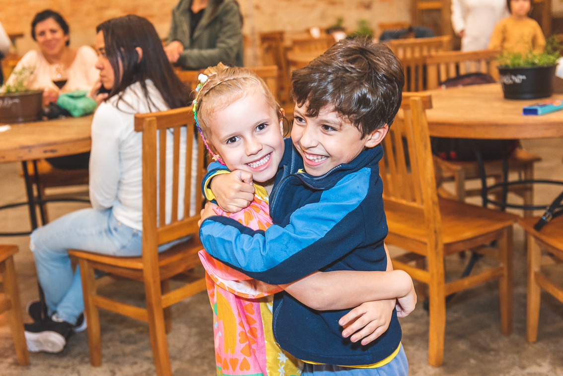 Duas crianças sorrindo e se abraçando com alegria durante uma festa infantil em ambiente interno, com mesas e convidados ao fundo.