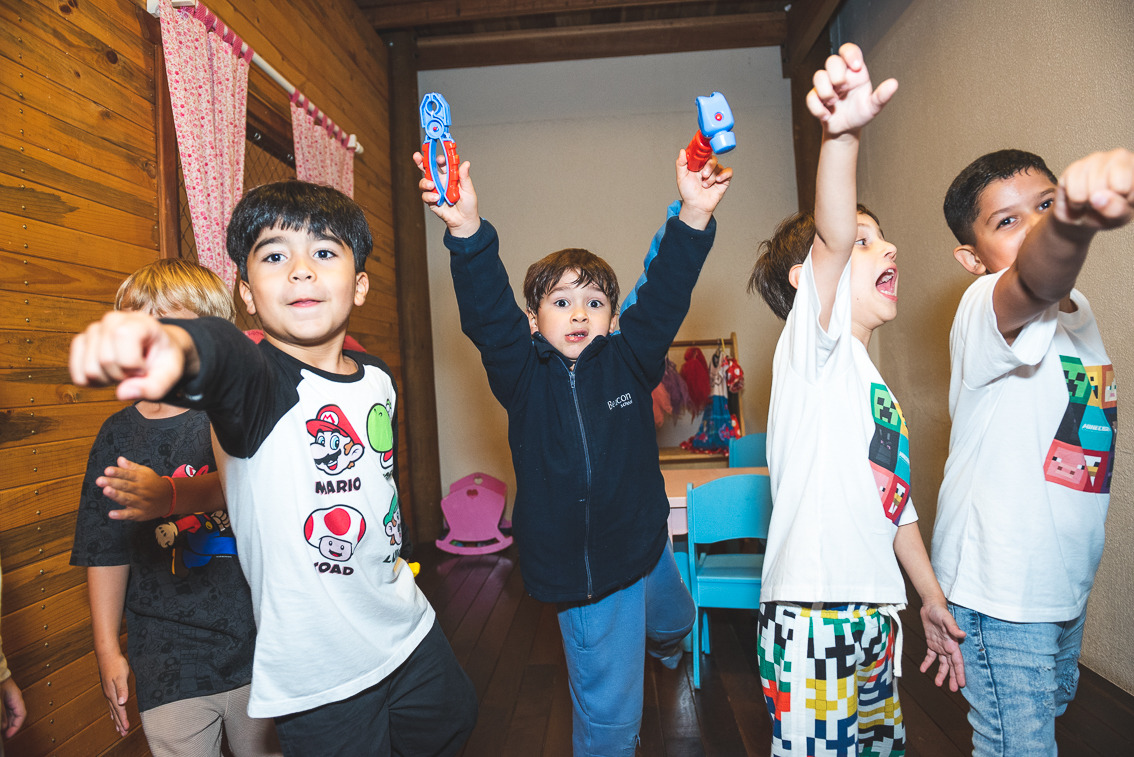 Grupo de meninos brincando animados durante uma festa infantil em ambiente interno, alguns levantando brinquedos e fazendo poses divertidas, em um espaço com paredes de madeira e decoração colorida ao fundo.
