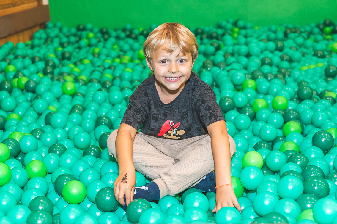 Menino sorrindo sentado em piscina de bolinhas verdes durante festa infantil, brincando e olhando para a câmera em ambiente de buffet infantil.