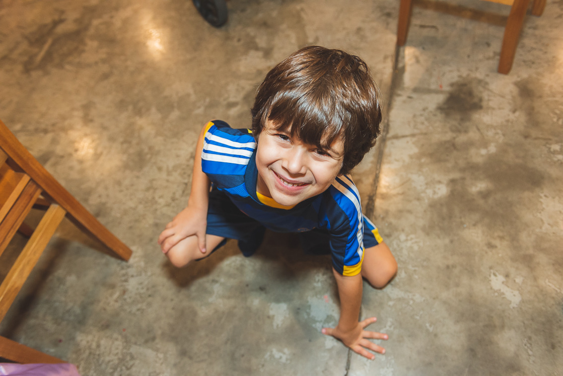 Menino sorrindo e brincando no chão durante festa infantil em buffet, olhando para a câmera em momento espontâneo de diversão.