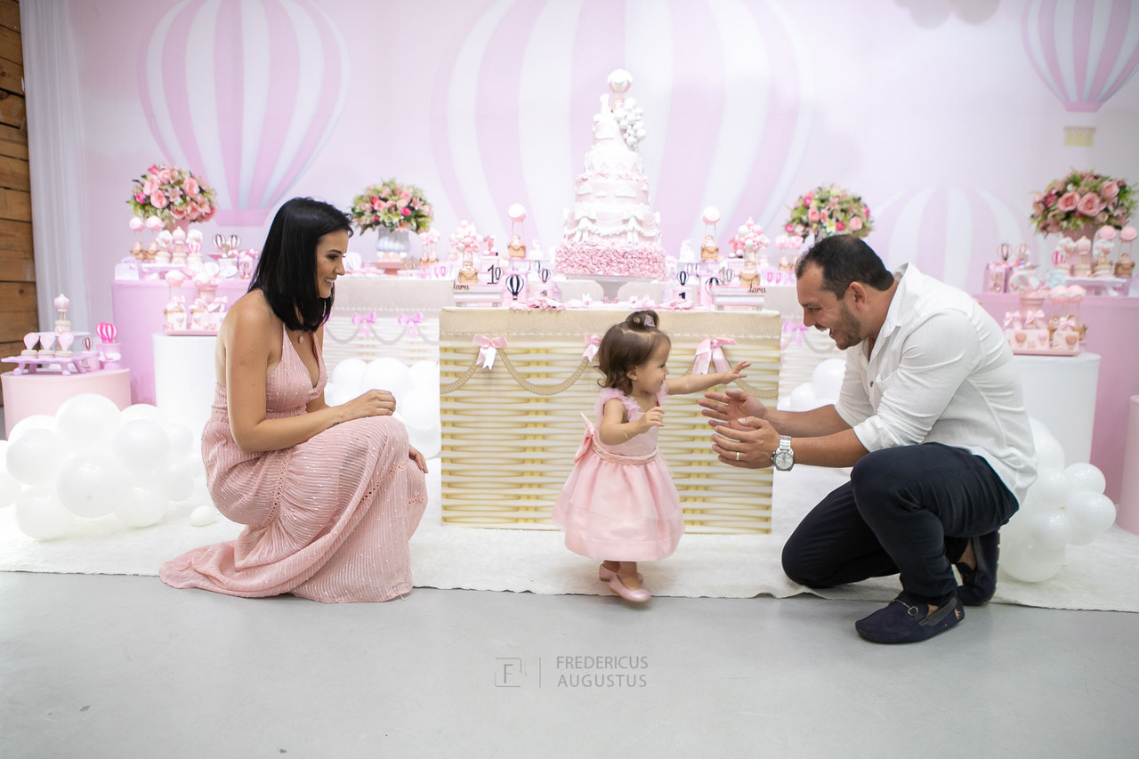 Foto da aniversariante na mesa do bolo com seus pais dando seus primeiros passos