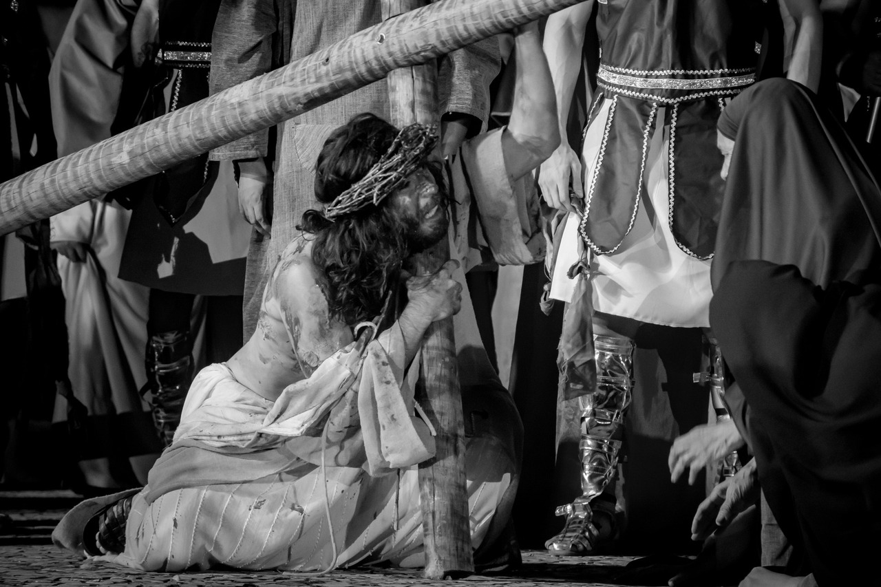 Fotografia em preto e branco de Jesus Cristo, com coroa de espinhos, carregando uma cruz pesada sobre o ombro, em meio à multidão, durante a encenação da Paixão de Cristo.