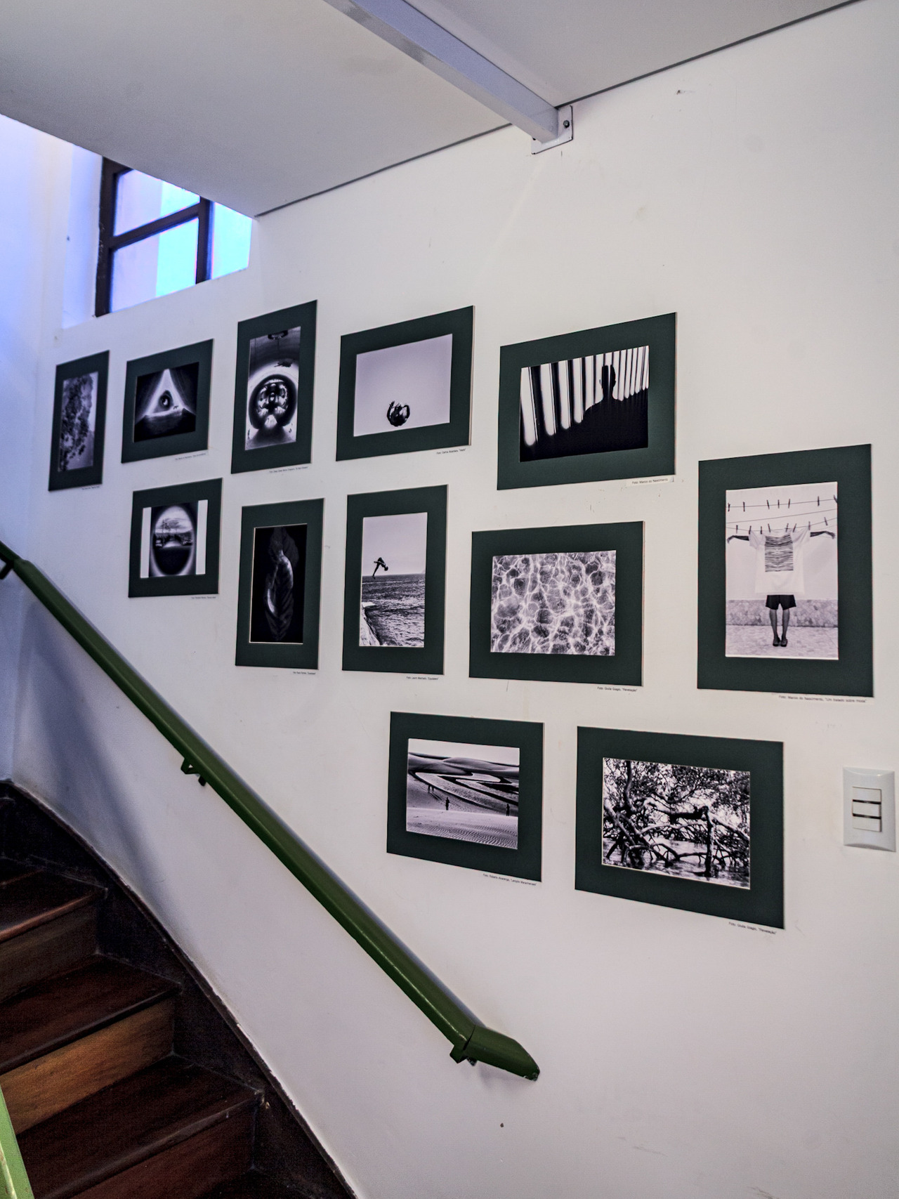Parede da escada com várias fotos em preto e branco Fotografias em preto e branco ocupam a parede da escada da galeria, com paspatur verde que une as obras de diferentes autores.
