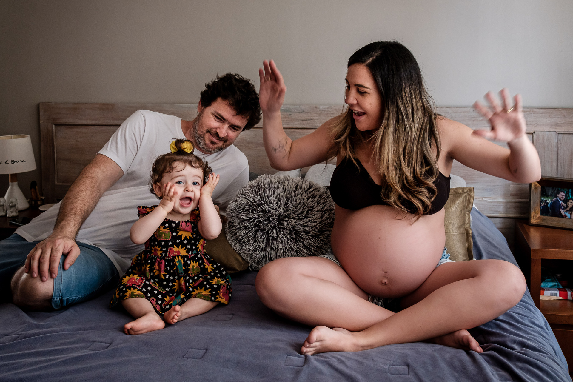 Família em momento espontâneo sobre a cama, com mãe grávida, pai e filha, em fotografia de Kelly Schmidt